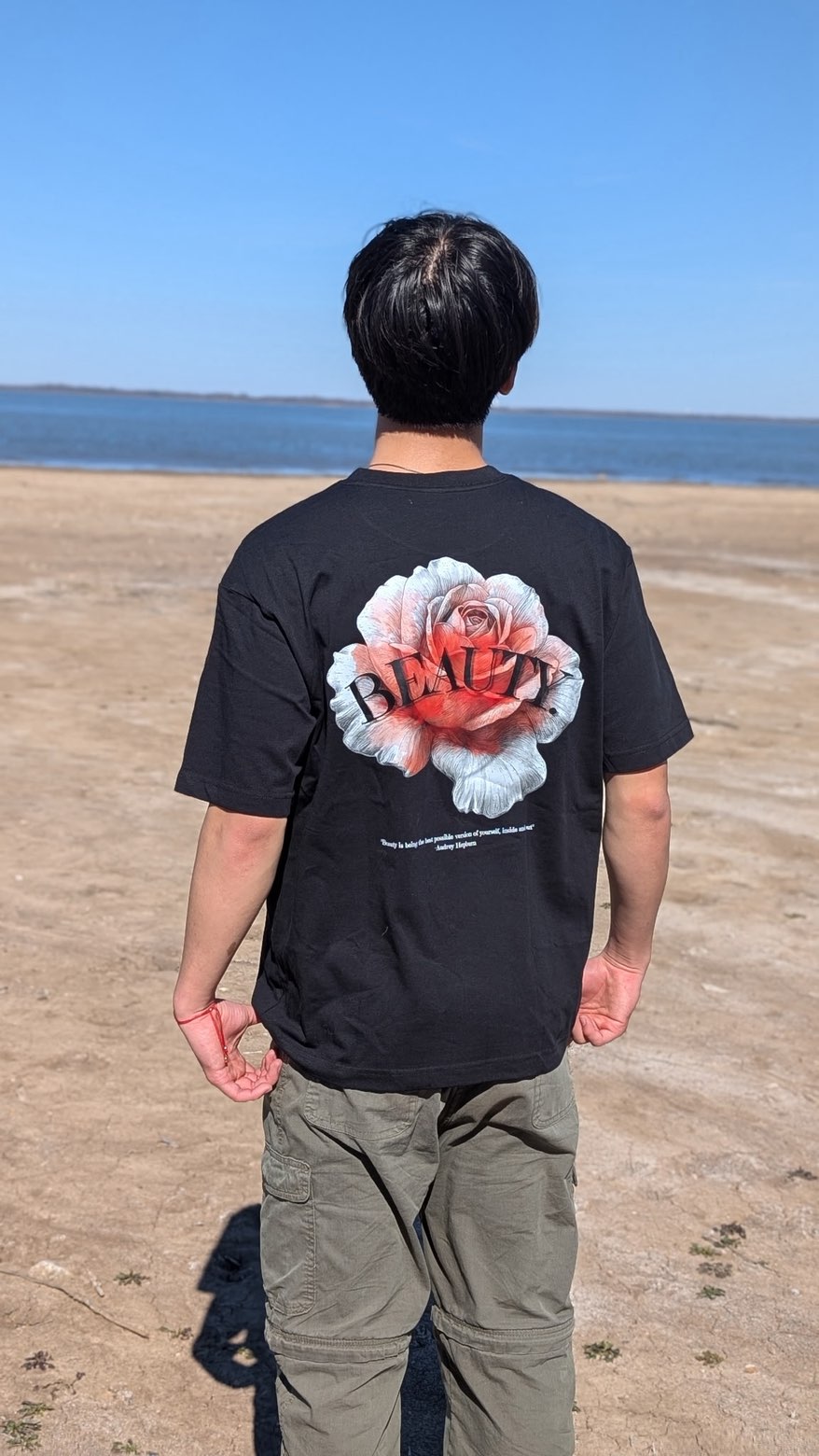 Back of the BEAUTY. Abnormal tee: black cotton with a red and white rose graphic, worn on a lakeside beach.