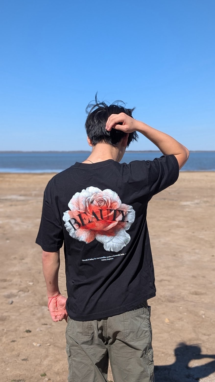 The same tee from behind, one hand in the hair, blue sky and water in the background.